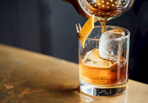 A whiskey glass on a bar counter with a large ice cube, a twist of orange zest, and a pour being topped from a bottle.