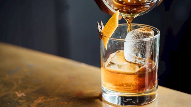 A whiskey glass on a bar counter with a large ice cube, a twist of orange zest, and a pour being topped from a bottle.