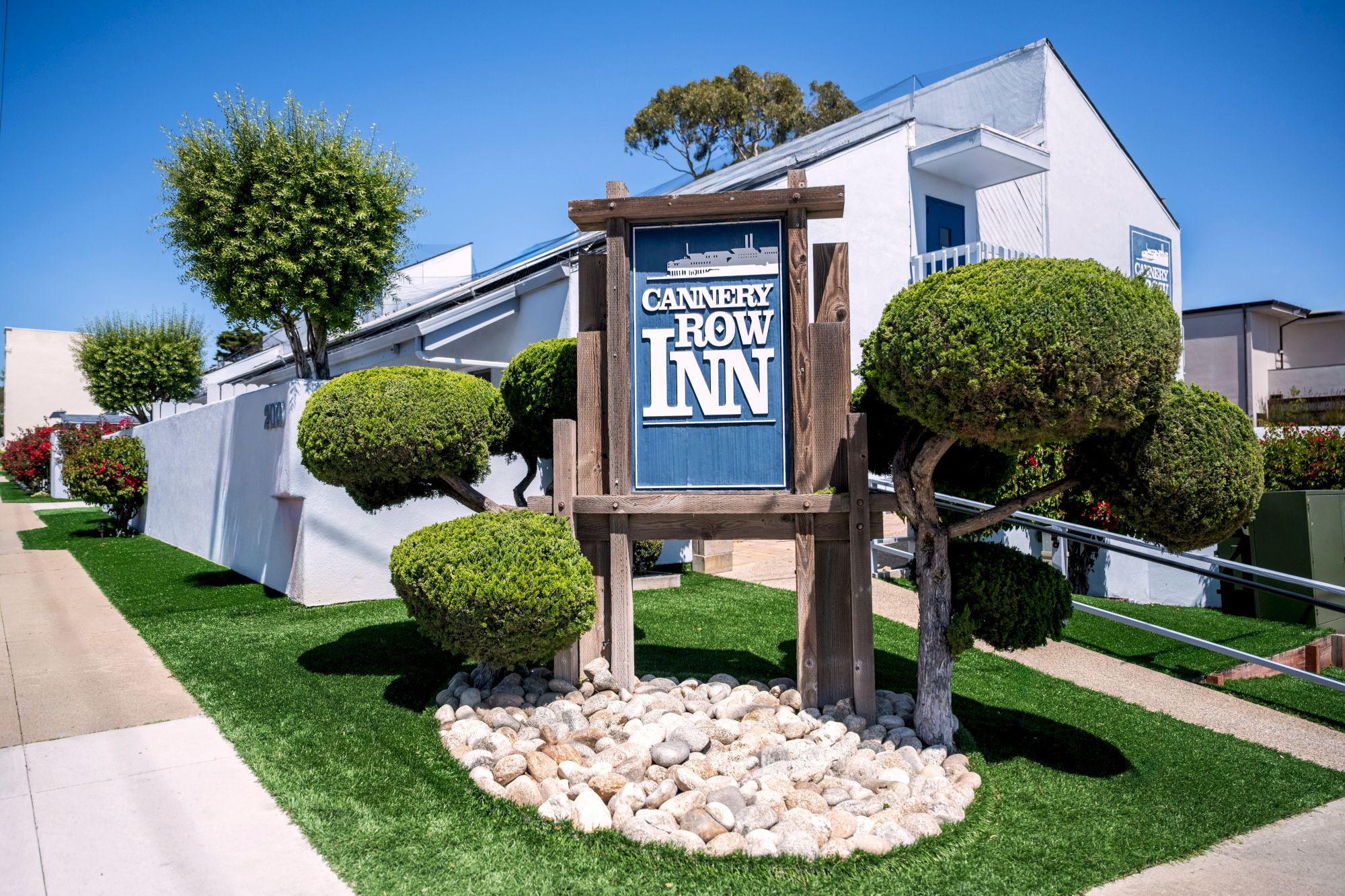 A small inn with a wooden sign reading &ldquo;Cannery Row Inn&rdquo; in front of a white, modern building surrounded by trimmed shrubs and rocks.