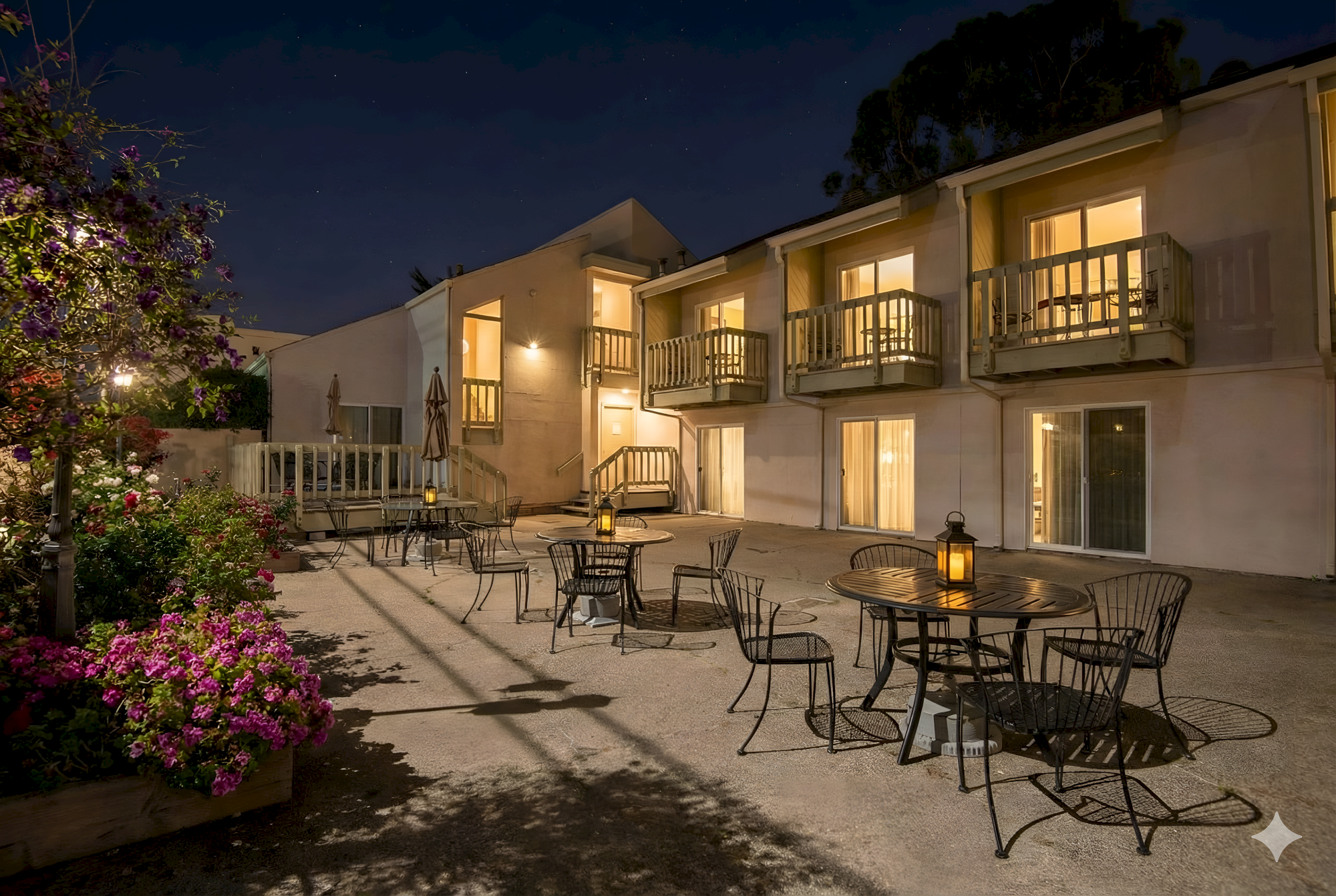 A quaint courtyard at night with several outdoor tables and chairs, lit building balconies, warm lights, and blooming flowers nearby.