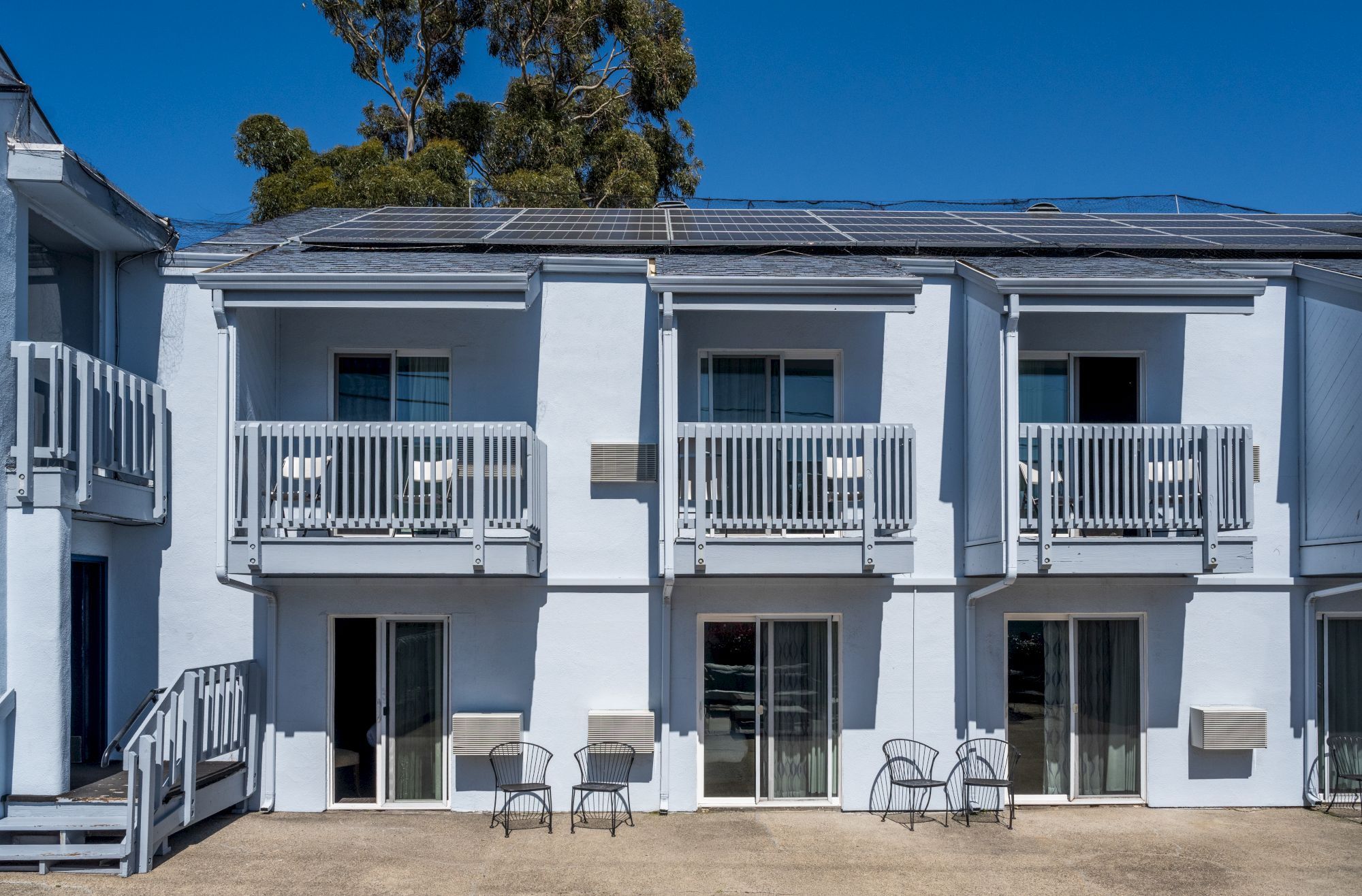 A two-story blue motel with white trim, balconies on the upper level, sliding glass doors, and a row of outdoor chairs on a sunny courtyard.