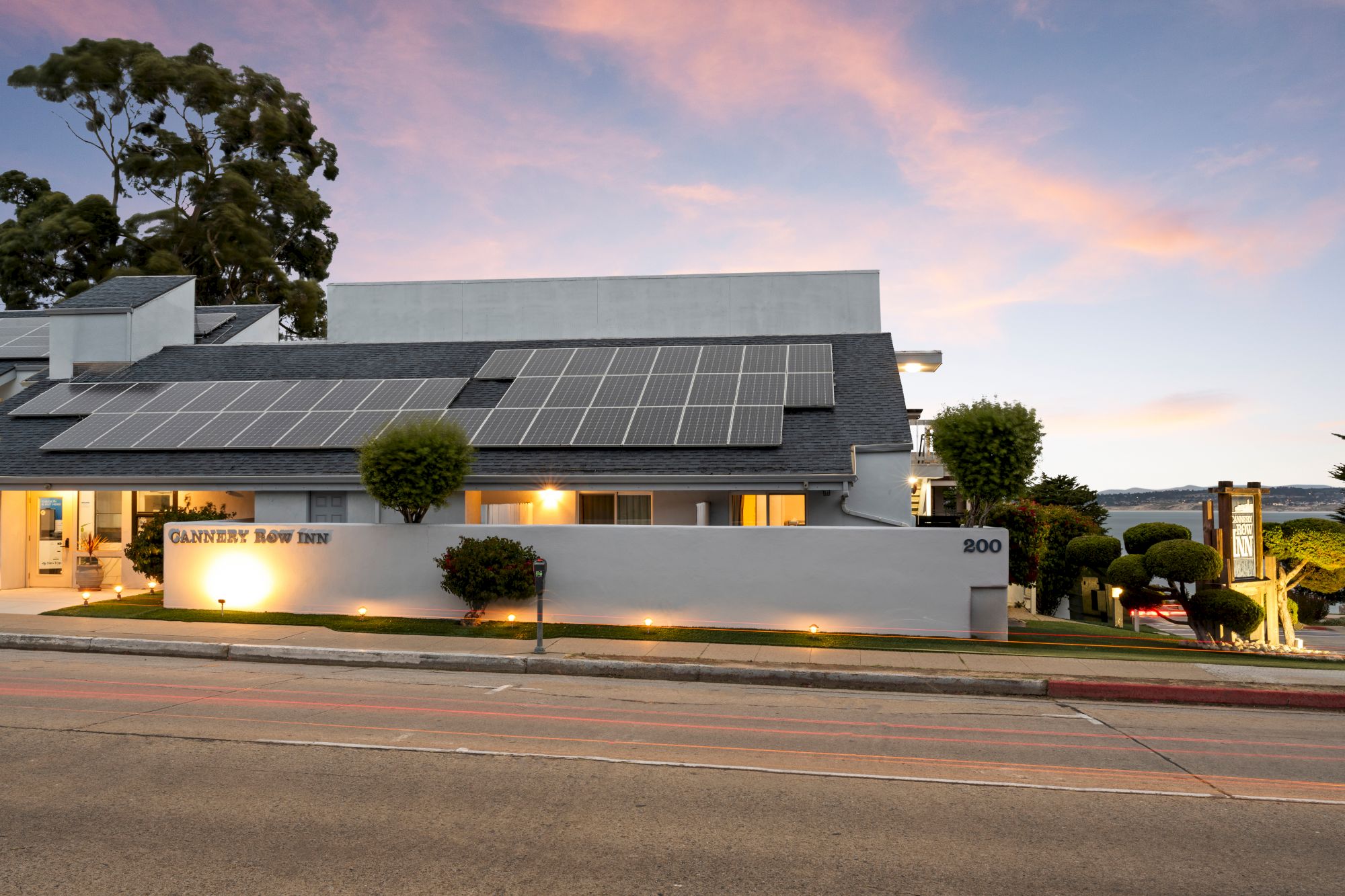 A modern white building with a row of solar panels on the roof, a low wall, manicured shrubs, and a calm sunset sky behind.