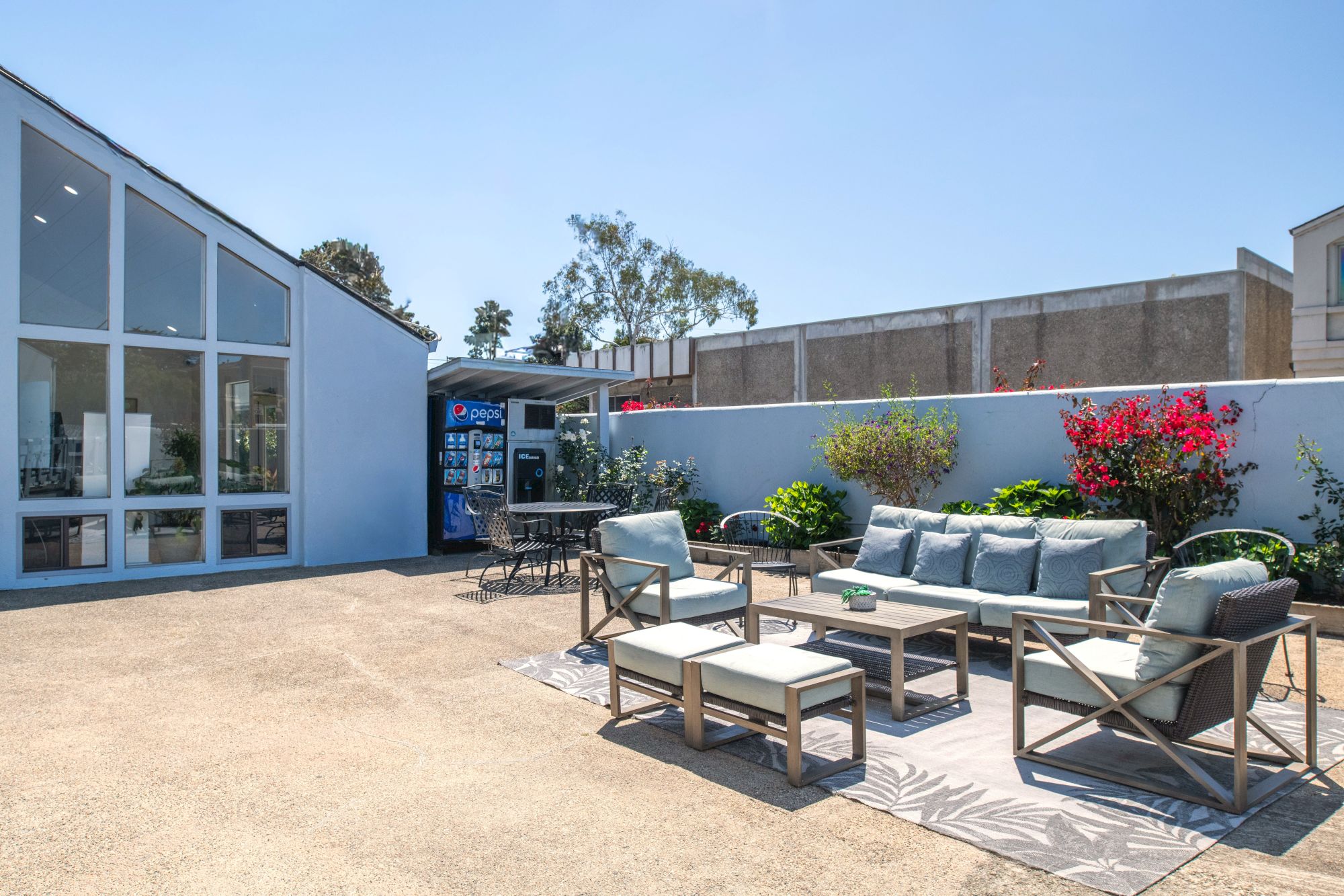 A modern outdoor patio with gray seating, a coffee table set, potted plants, and a blue building with large glass doors under a clear sky.