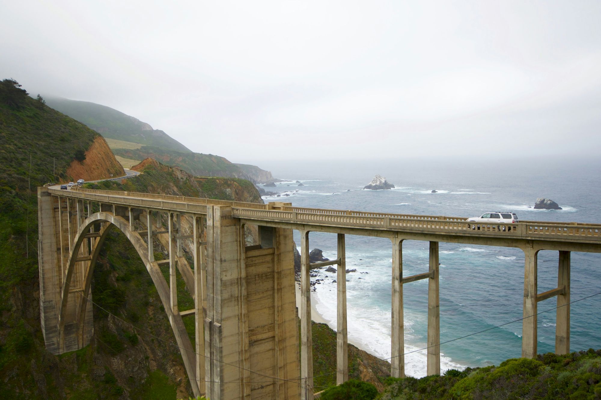 A tall concrete arch bridge spans a rugged coastal cliff, with the ocean below and rocky shores in the distance along a misty coastline.