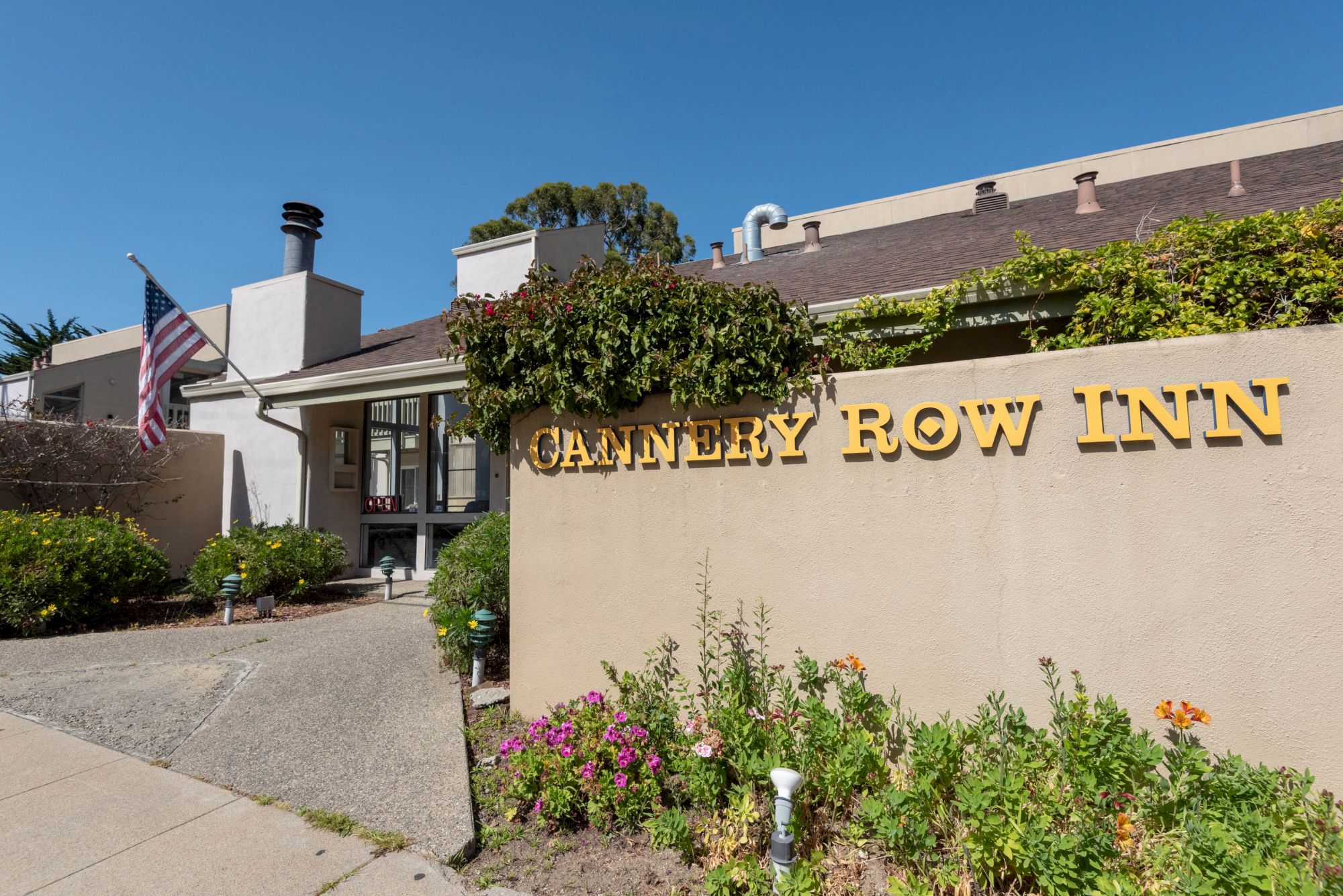 A motel entrance with a beige sign reading &ldquo;CANTERY ROW INN&rdquo; (likely "Cantlery"?) and a small garden in front; American flag on a pole by the doorway, clear blue sky.