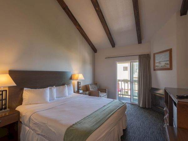 A hotel room with two large beds, dark headboard, lamps, a small seating area, balcony door, and neutral decor with wooden ceiling beams.