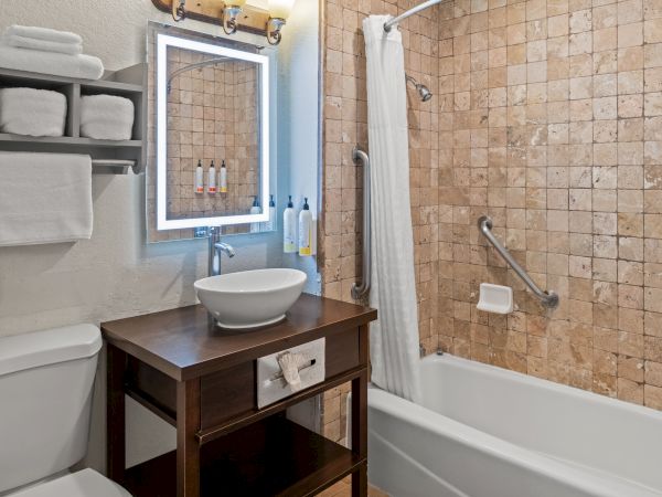 A compact bathroom with a wooden vanity, vessel sink, wall mirror, open shelving, and a tiled bathtub/shower combo.