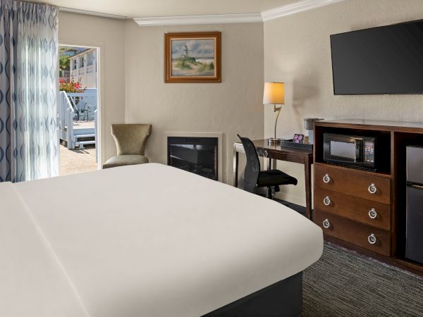 A hotel room with a large bed, a dresser with a TV, a mini-fridge, a desk and chair, a lamp, and a small seating area near a doorway with blue curtains.