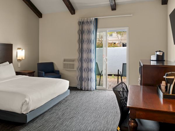 A hotel room with a large bed, desk, chair, and a sliding door to a balcony; neutral tones, exposed ceiling beams, and a TV on the wall.