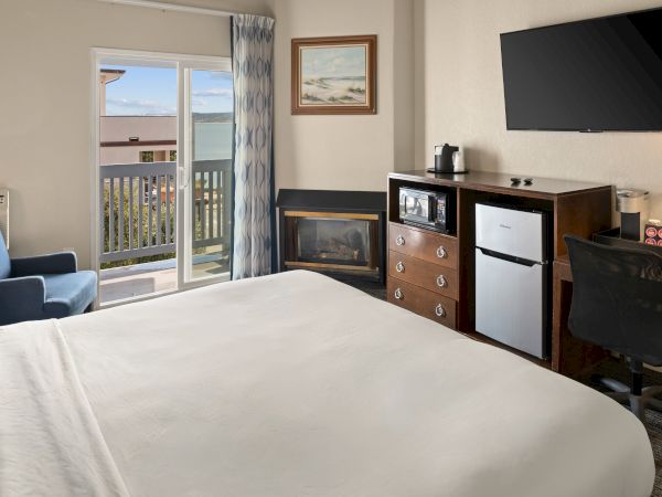 A bright hotel room with a neatly made bed, a desk, a flat-screen TV, mini-fridge, fireplace, and a balcony view through glass doors.