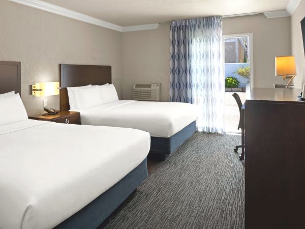 Hotel room with two queen beds, nightstands, lamps, a desk, chair, and sheer curtains by a window; neutral tones and contemporary decor.