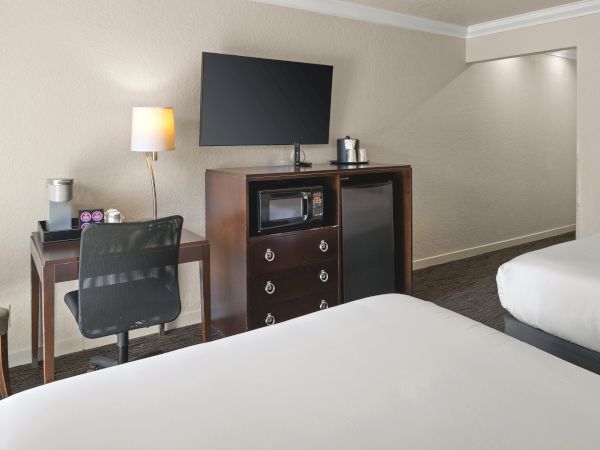 A clean hotel room with two beds, a desk and chair, a wall-mounted TV, a mini-fridge/hutch, and soft lighting.