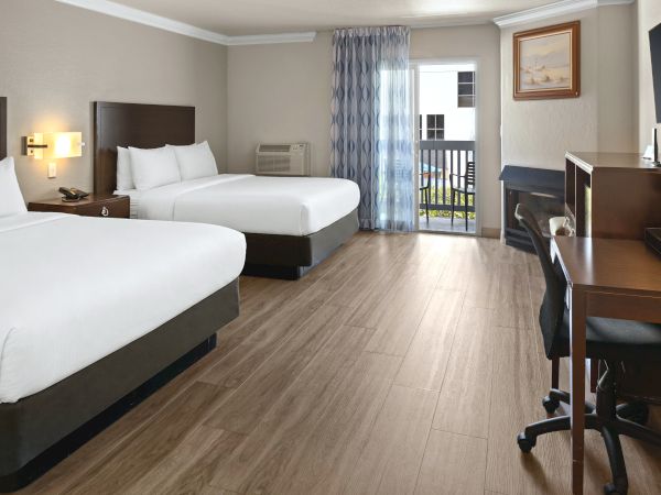 Two hotel beds, a desk and chair, and a door to a balcony in a bright, modern room with light wood floors.