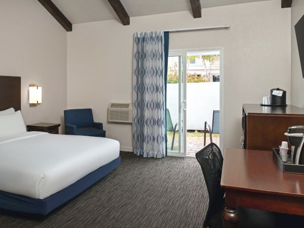 A modern hotel room with a king bed, a blue chair, a desk, and a sliding door to a small balcony; neutral tones and exposed ceiling beams.