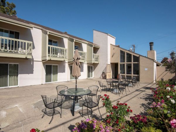 A sunny motel-style courtyard with white two-story units, patio tables and chairs, an umbrella, and blooming flowers along a concrete walkway.