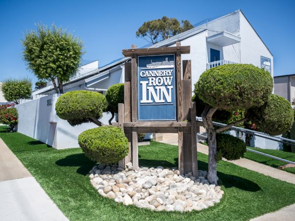 A neat roadside scene of a white building with a blue-tiled roof, manicured shrubs, and a wooden sign that reads &ldquo;Cannery Row Inn.&rdquo;