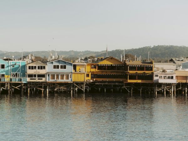 Colorful beachside huts on stilts along a calm harbor, painted in pastel blues, yellows, and browns, reflecting softly in the water.
