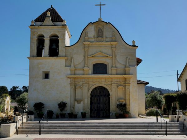 A church with a central ornate entrance, a cross on top, and a bell tower on the left; steps lead up to a large wooden door.