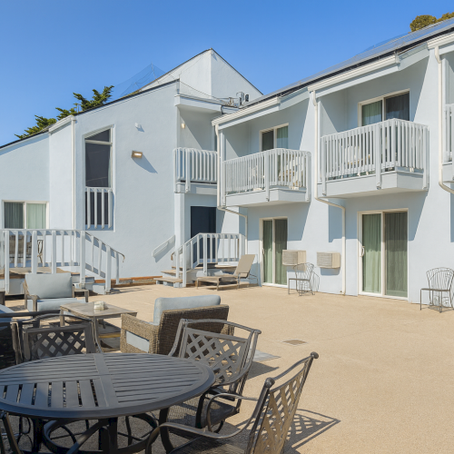 A sunny coastal row of light blue and white townhomes with balconies, an outdoor seating area, and parked cars along a narrow street.