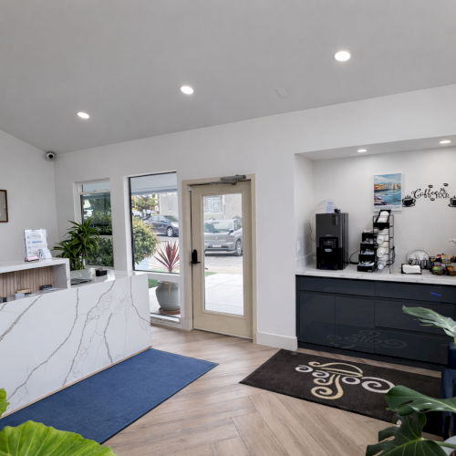 A modern reception area with a marble-front desk, black and white accents, plants, a coffee/snack station, and a glass door entrance.