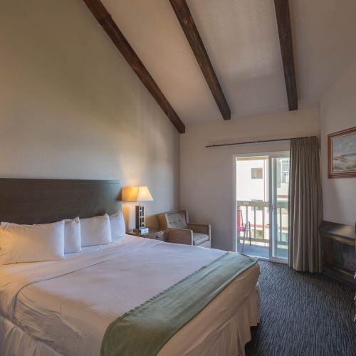 A cozy hotel room with two queen beds, warm lamps, exposed wooden beams, a balcony door, and a desk with a chair.