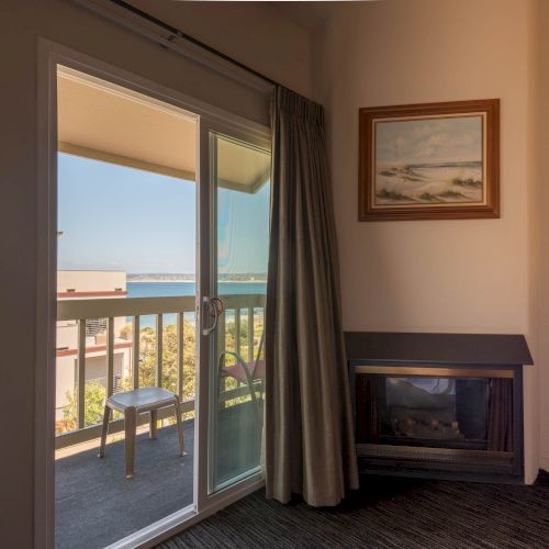 A hotel room with a sliding door opening to a balcony view of the sea; there&rsquo;s a small table, a chair on the balcony, a framed picture, and a fireplace in the corner, ending with the sea breeze.