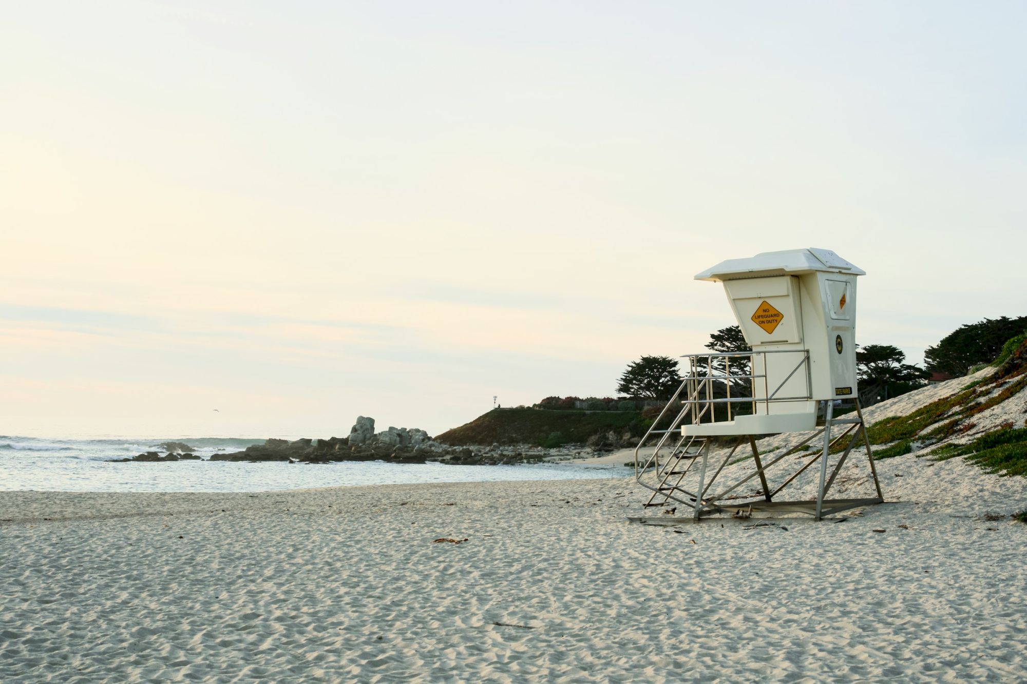A quiet beach with a lifeguard tower on the sandy shore, gentle waves, and a pale sky at either dawn or dusk.