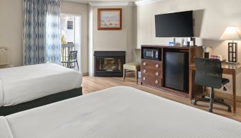 Hotel room with two white beds, a wood desk and chair, a wall-mounted TV, a dresser with a mini-fridge, a small fireplace, and a balcony door with blue patterned curtains.