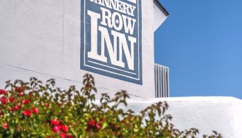A white-walled building with a blue &ldquo;CANNERY ROW INN&rdquo; sign mounted high on the wall, and a flowering shrub in the foreground under a clear blue sky.
