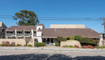 A single-story suburban building with a long, low roofline, beige walls, small front yard shrubs, a sidewalk, and clear blue sky above.