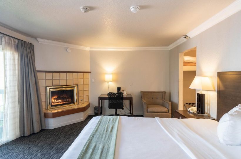 A hotel room with a bed in the foreground, a lit fireplace on the left, a small desk and chair in the middle, and a seating area with lamps on the right.
