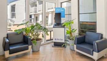 A modern waiting area with two navy chairs, plants, and a check-in kiosk by tall windows. ends.