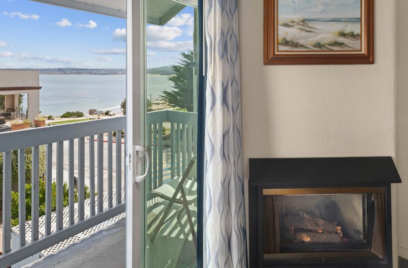 A hotel room with a sliding glass door to a balcony overlooking the ocean, a faux fireplace, and a framed beach painting on the wall.