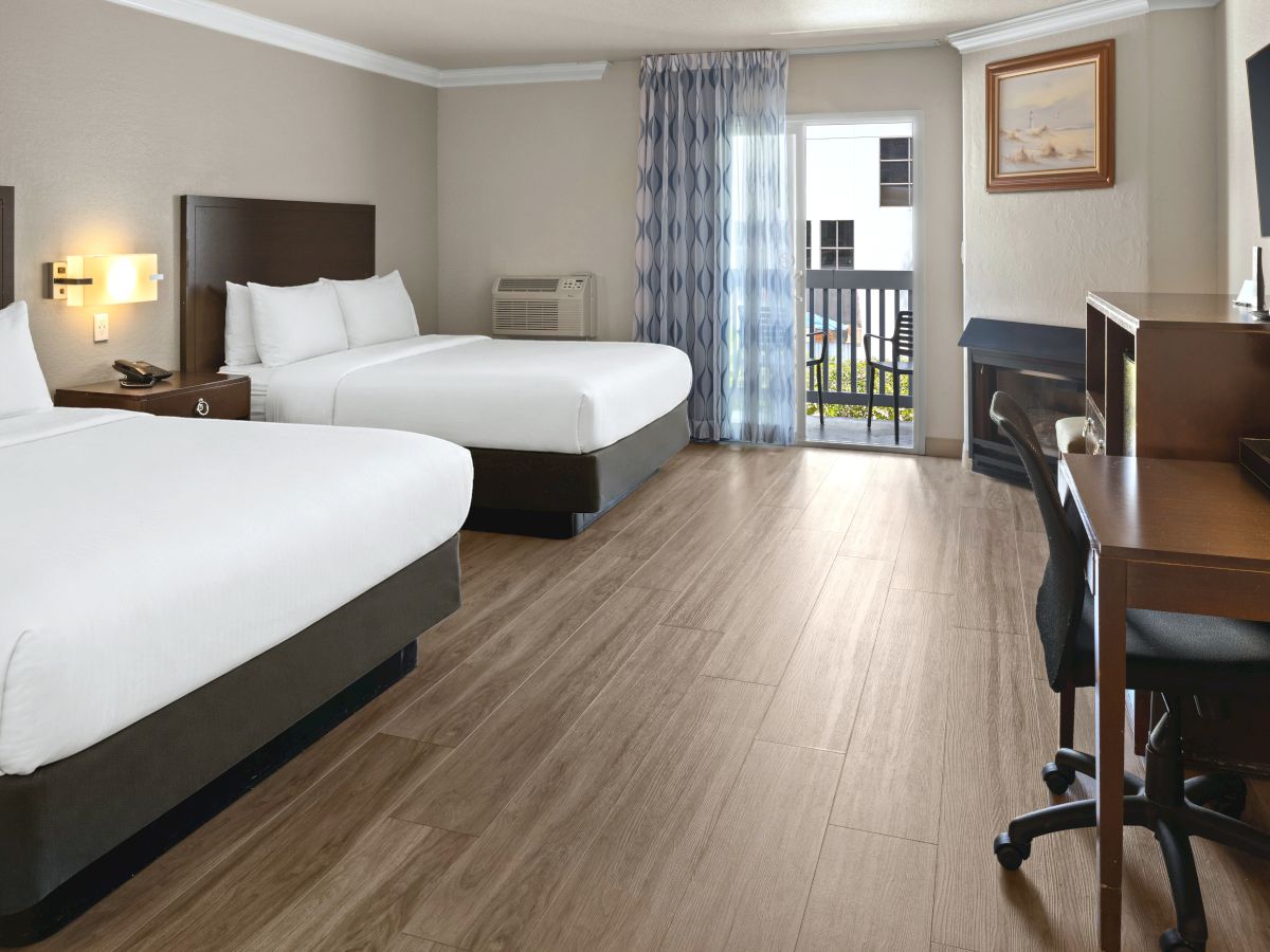 A hotel room with two queen beds, wooden floor, desk and chair, a balcony with sheer blue curtains, and a dresser with a TV, all bright and neat.