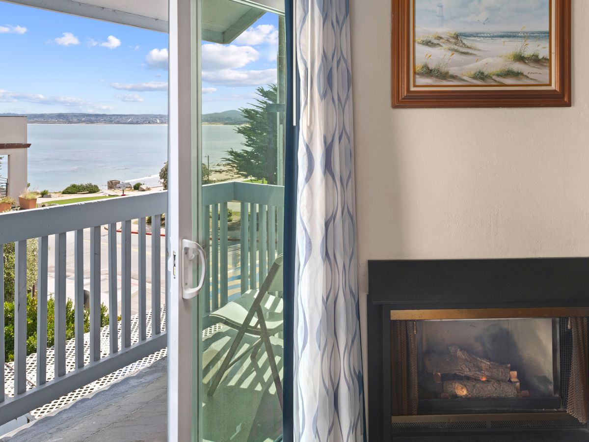 A cozy balcony room with a glass door opening to a sunny ocean view, a railing outside, patterned curtains, and a modern fireplace beside a framed painting.