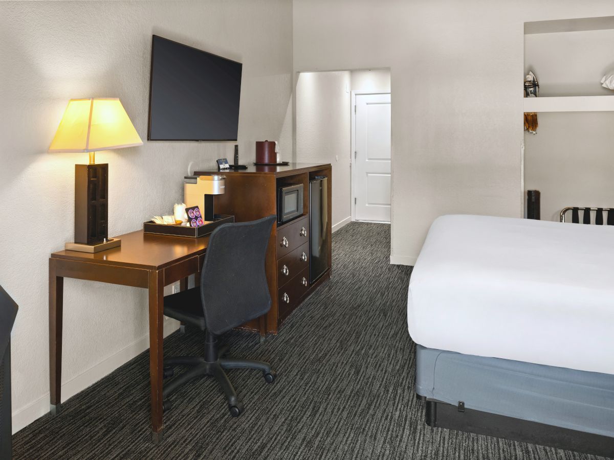 A hotel room with a desk, chair, TV, dresser, lamp, and a neatly made bed in the foreground, plus a door and open shelves in the background.