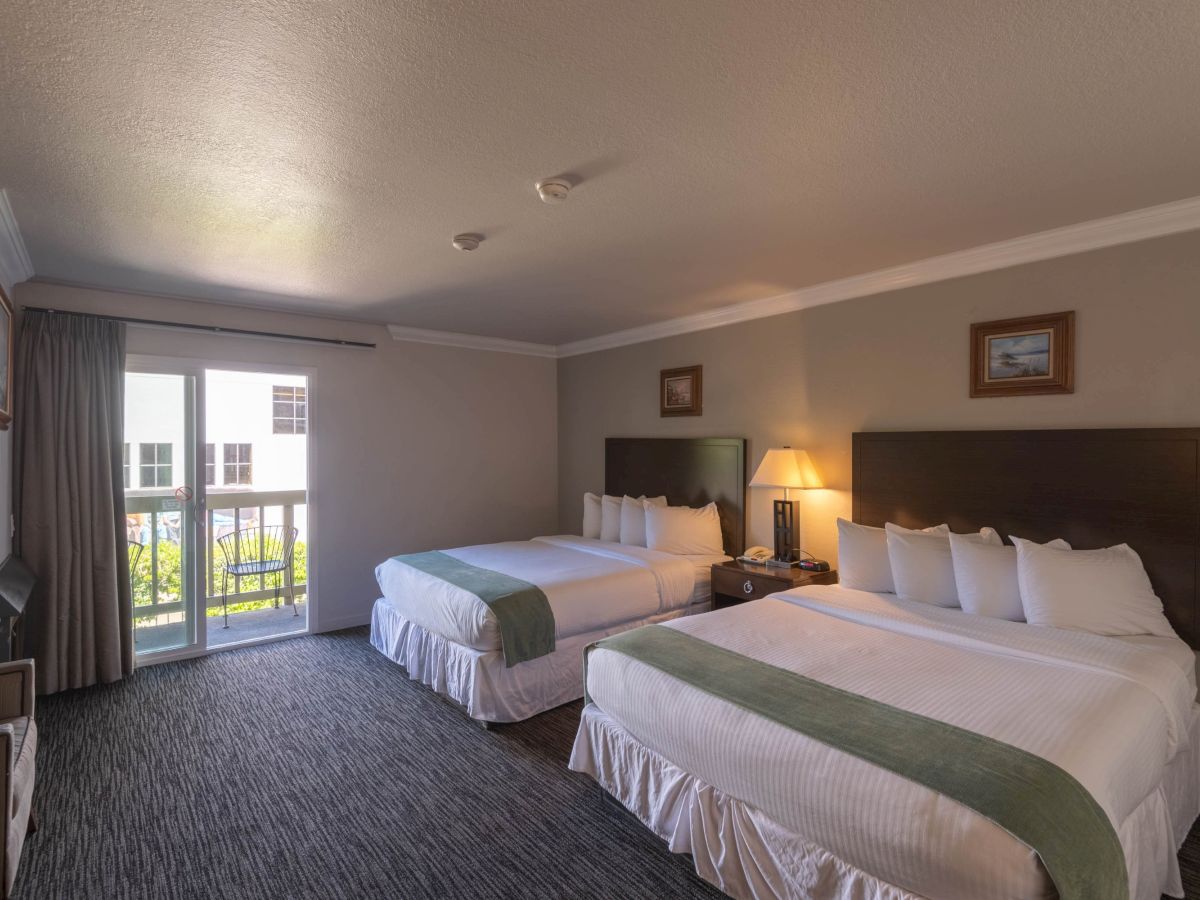 A hotel room with two queen beds, grey walls, nightstand, lamps, and a balcony door with curtains, overlooking a courtyard.