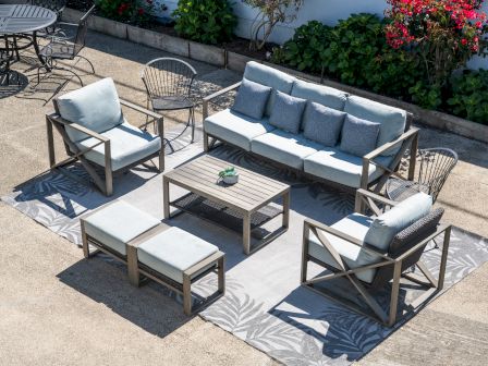 Outdoor patio furniture set with light-gray cushions on a wood frame, including a sofa, two armchairs, two ottomans, a coffee table, and side tables on a patterned rug. Top at 140 characters, always ending the sentence.