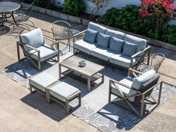 Outdoor patio furniture set with light-gray cushions on a wood frame, including a sofa, two armchairs, two ottomans, a coffee table, and side tables on a patterned rug. Top at 140 characters, always ending the sentence.
