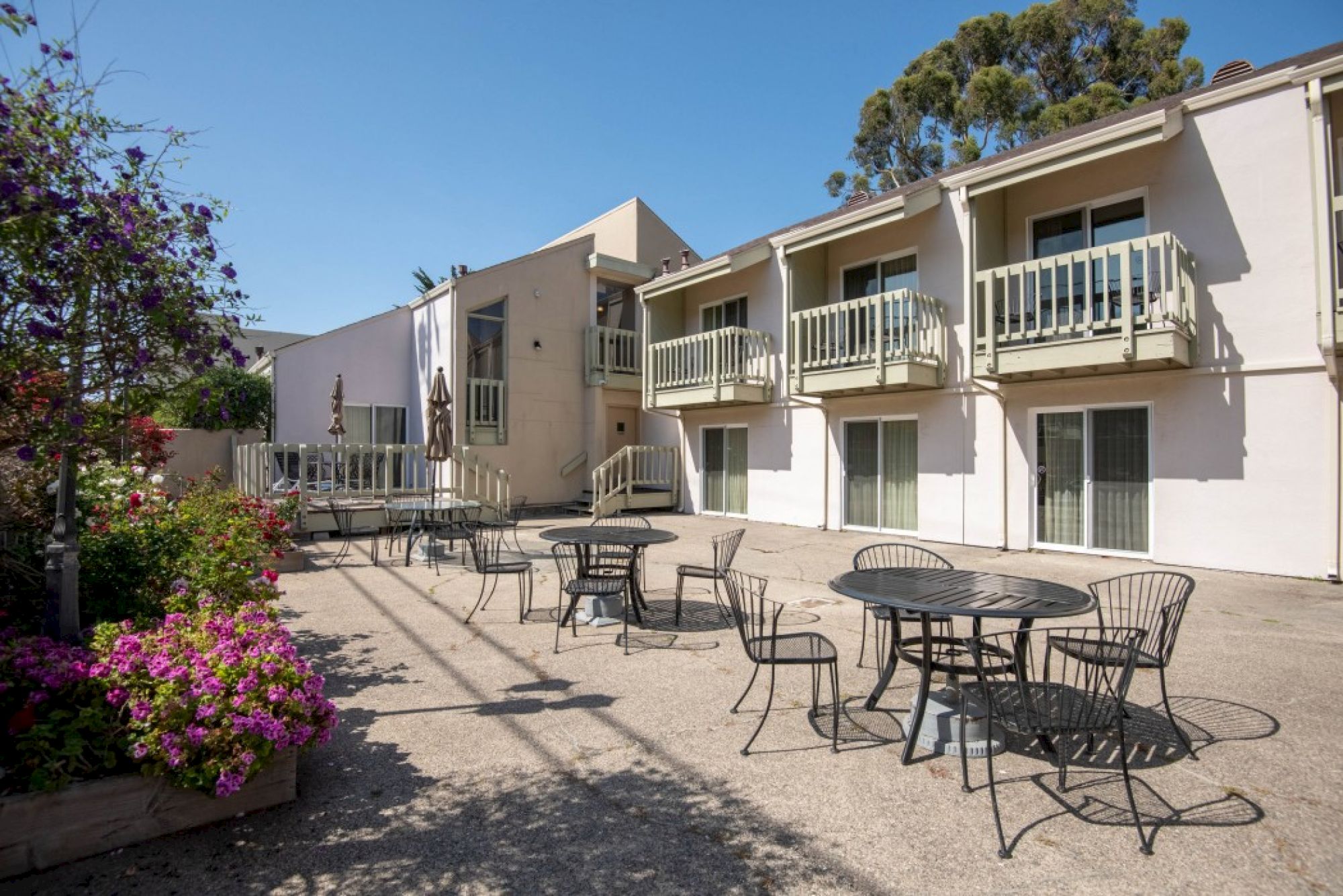 A sunny motel-style courtyard with metal tables and chairs, a two-story beige building, balconies, and flowering plants along the path.
