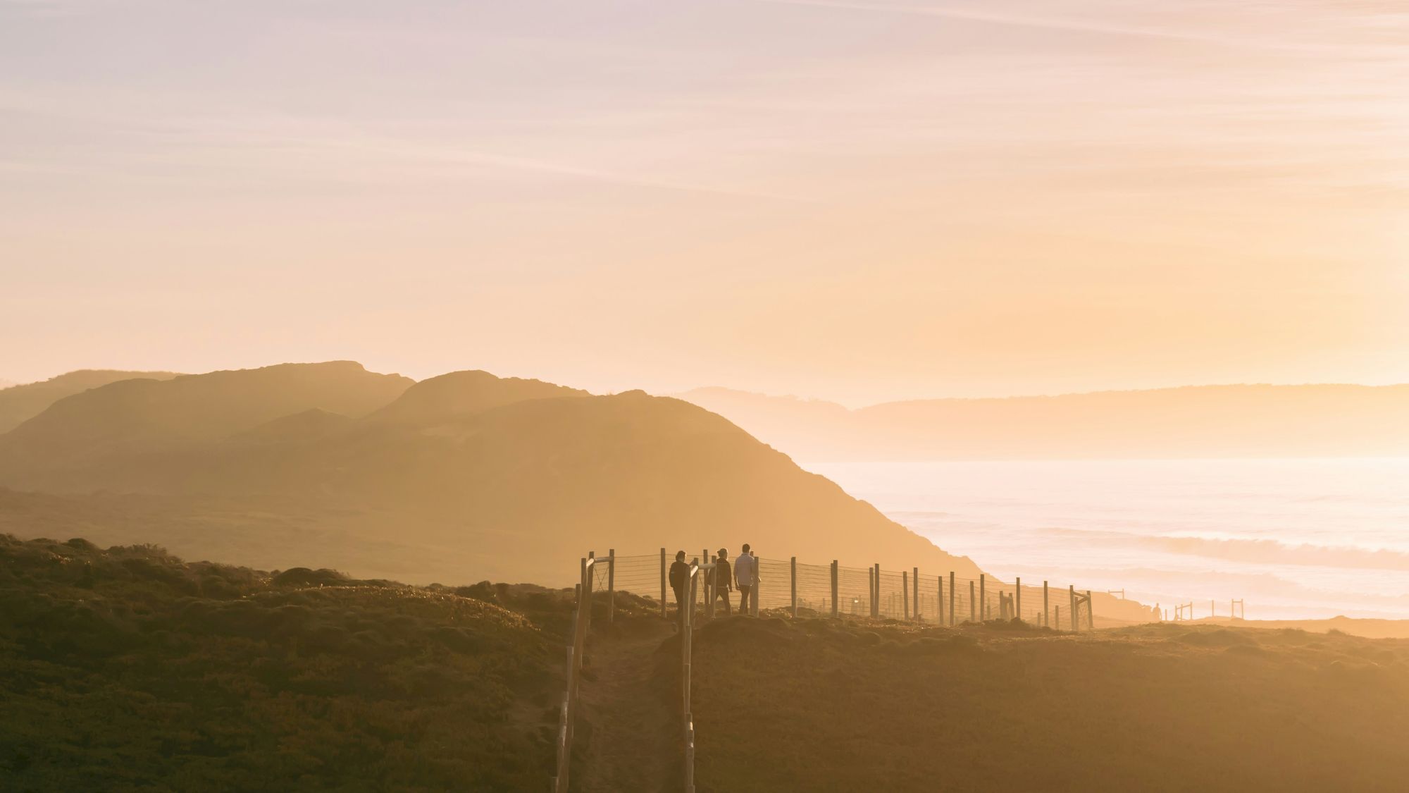 A group of people stand near a fence along a cliff path at sunset, with hazy mountains and the sea glow behind them.