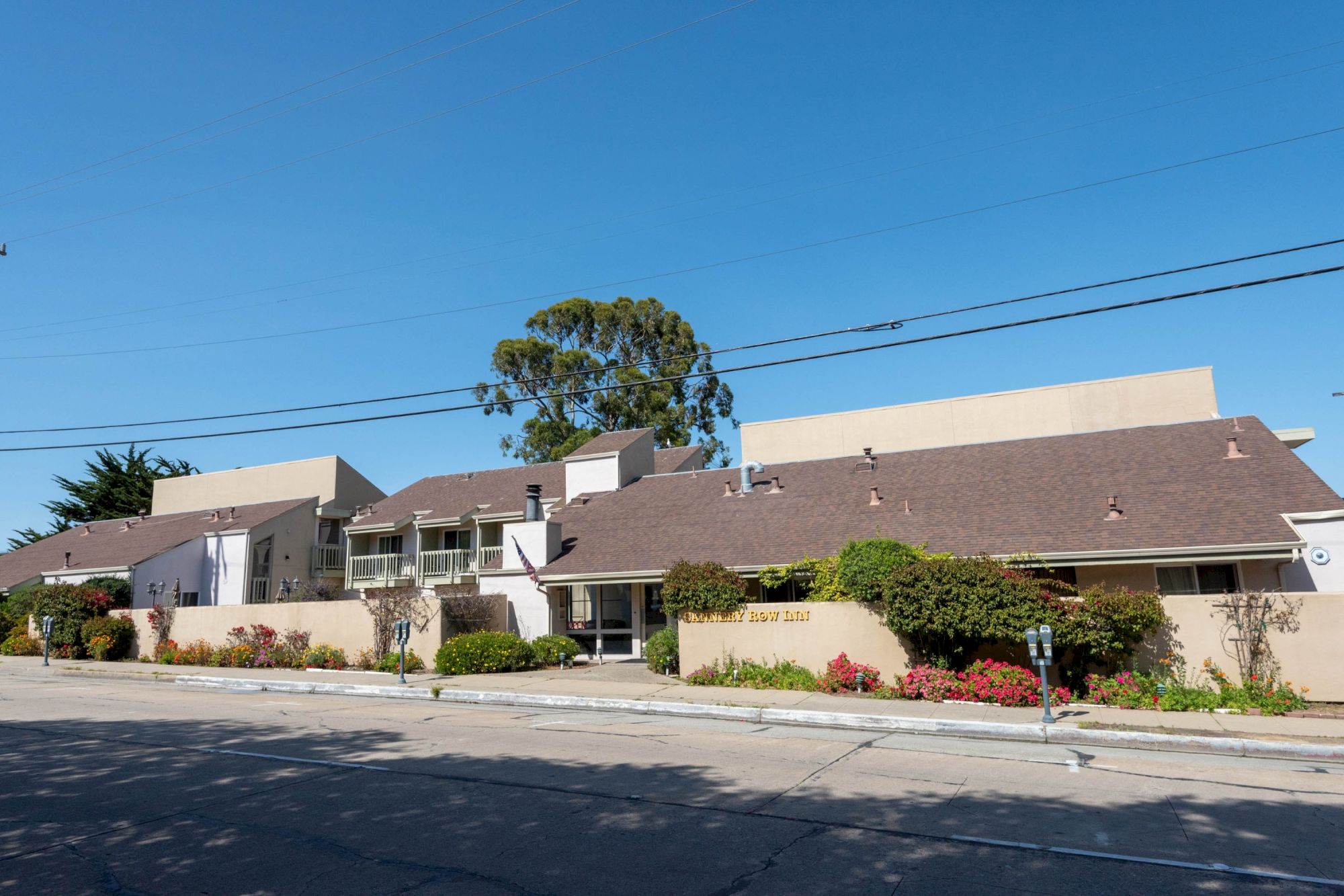 A row of single-story suburban apartment buildings with beige walls, brown roofs, trimmed shrubs, and a clear blue sky behind them.