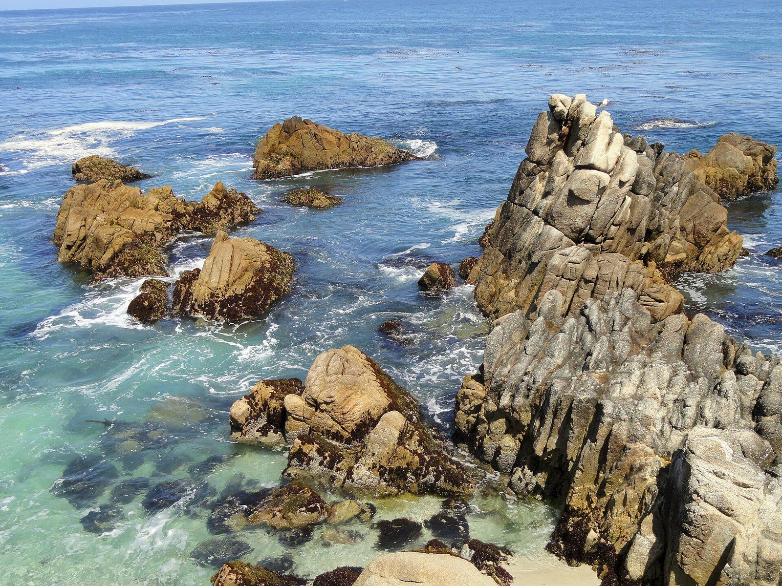 Rocky coastline with jagged granite outcrops as waves crash into the blue sea under a clear horizon.