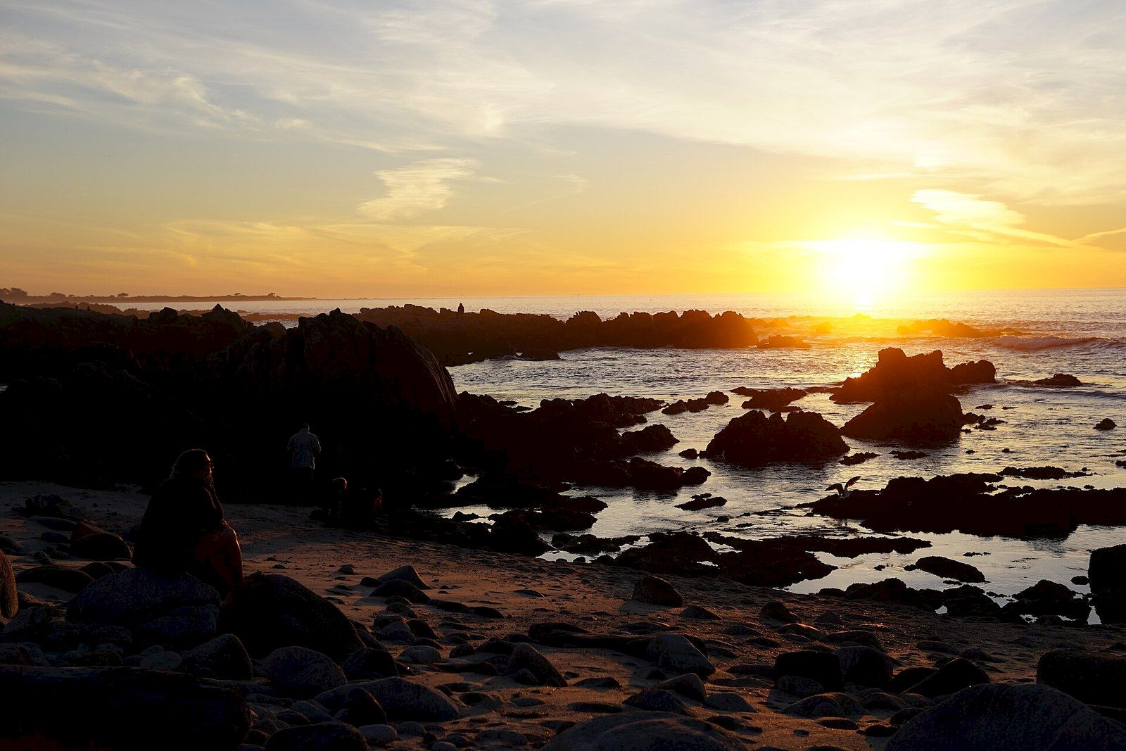 A peaceful rocky beach at sunset with waves, scattered boulders, and a couple watching the sun sink over the ocean.