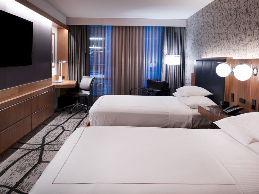 A modern hotel room with two neatly made beds, a desk, TV, and soft lighting by the windows.