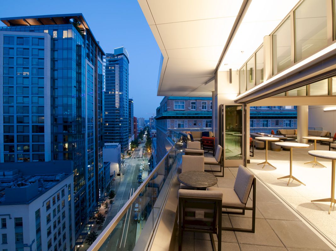 Modern high-rise balcony with outdoor seating overlooks a downtown street between skyscrapers at dusk.
